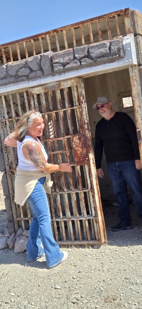 A man and a woman stand next to an old, rusty jail cell door. The woman smiles and touches the door.