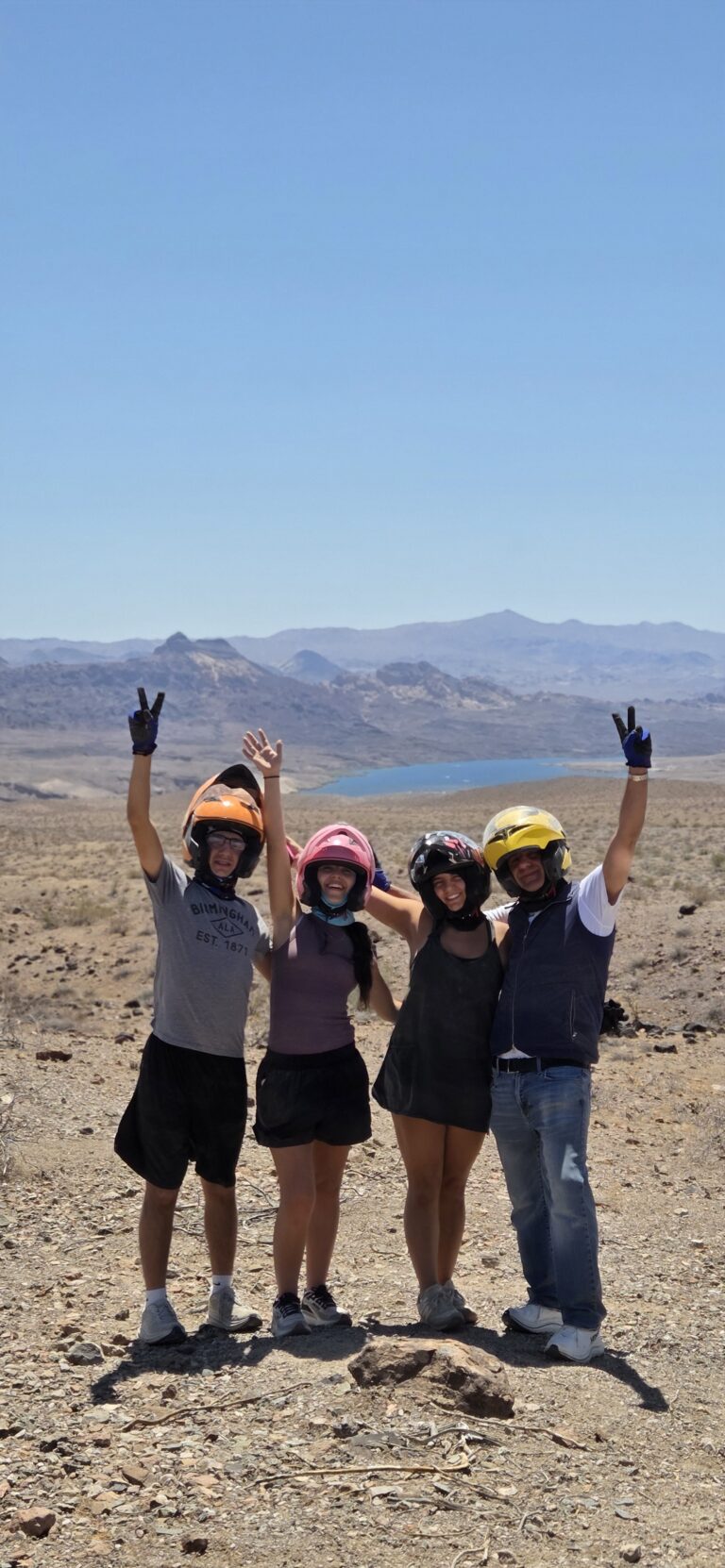 Four people in helmets stand in a desert landscape, waving at the camera with a lake and mountains in the background.