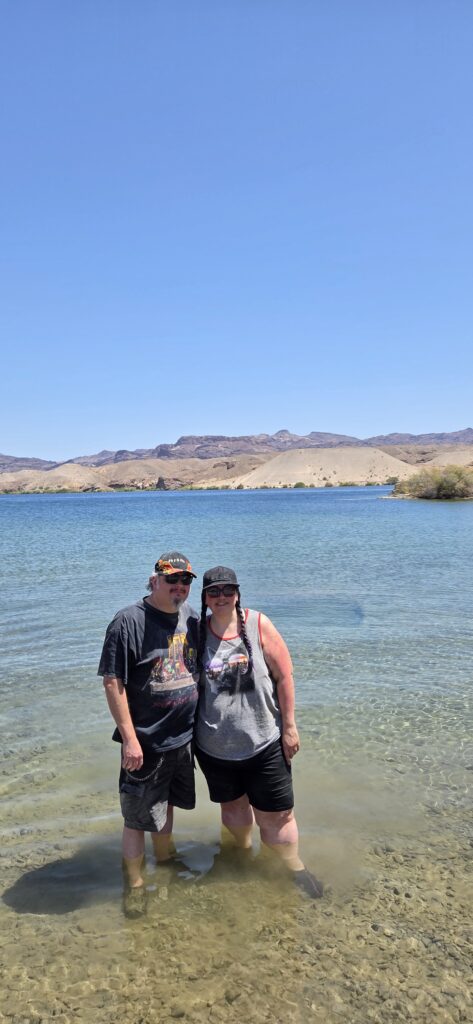 Two people enjoy the clear Colorado River near Las Vegas, Nevada, smiling in casual clothes and hats under a bright blue sky.