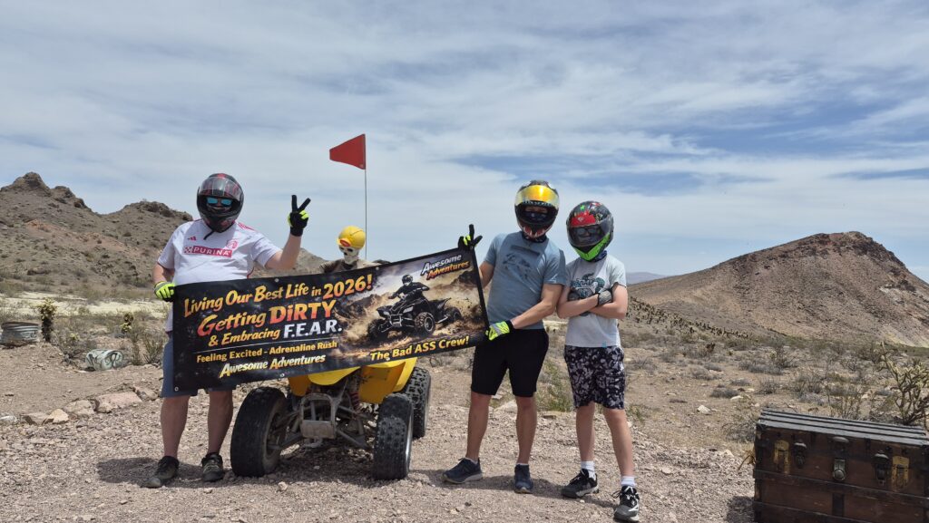Group in helmets poses with peace signs by yellow ATV on Nevada rocky terrain, banner reads “Living Our Best Life in 2026!”.