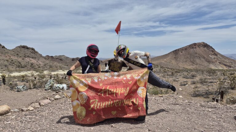 Two people in helmets hold a “Happy Anniversary” banner during an ATV tour near Las Vegas, Nevada, with scenic desert and mountains.