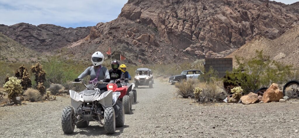 ATV riders explore a rocky Nevada desert trail near Las Vegas with old cars, mountains, and RZR off-road tours under a sunny sky.