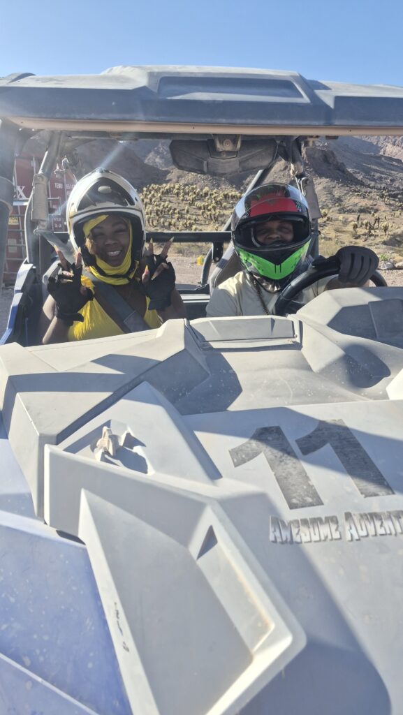 Smiling riders in helmets enjoy an ATV tour near Las Vegas, Nevada, flashing peace signs amid rocky desert terrain.
