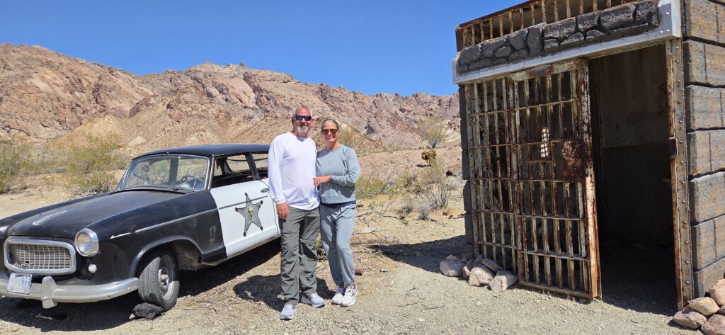 Smiling couple by vintage police car and rustic jail in Nevada desert near Las Vegas—perfect for Ghost Town sightseeing tours.