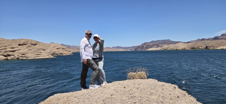 Two people stand on a rocky Nevada shore by the Colorado River near Las Vegas, mountains and blue sky in the background.