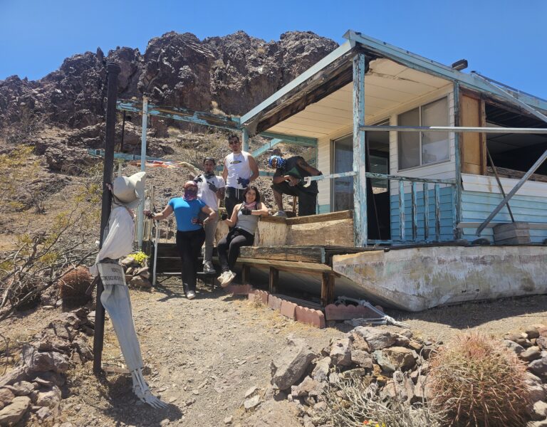 Six people smile on ghost town steps near Las Vegas, Nevada, with a scarecrow, cacti, and desert plants in the background.