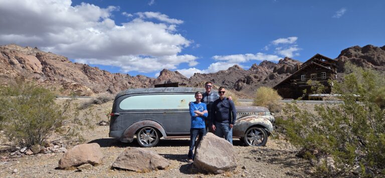 Three people pose by a rusty vintage car in a Nevada desert ghost town near Las Vegas, mountains and old wooden building behind.