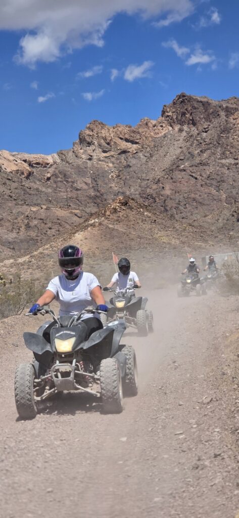 ATV riders in helmets and white shirts explore a dusty Nevada trail near Las Vegas with mountains, blue sky, and rising dust.