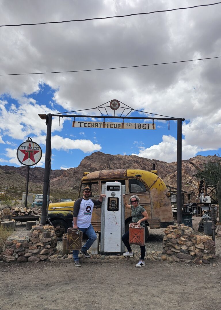 Two people pose by a vintage gas pump and yellow bus at Techatticup Ghost Town near Las Vegas, Nevada—great for ATV tours.
