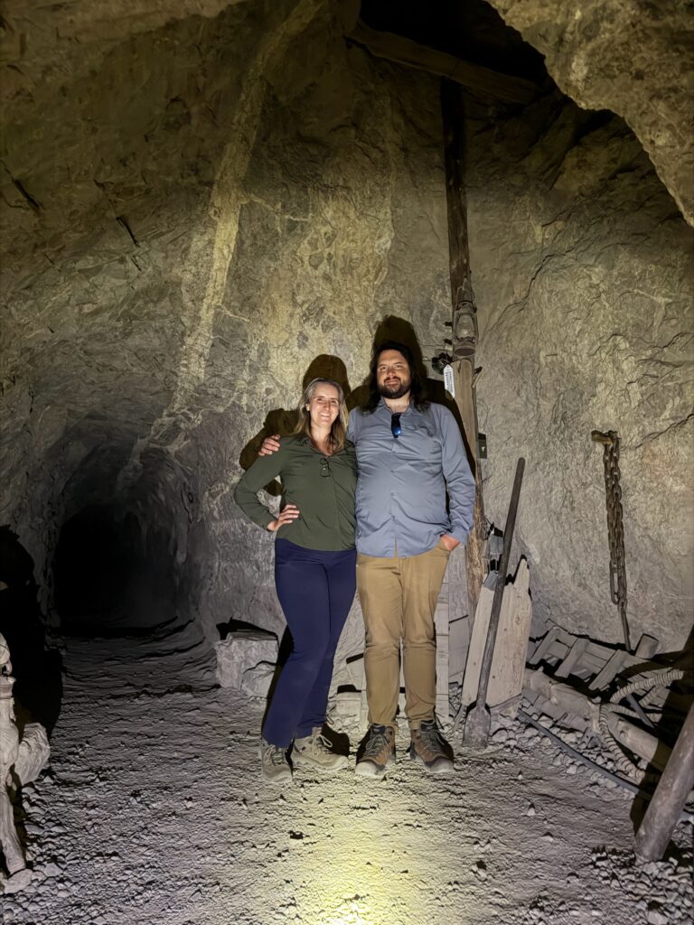 Two visitors smile in a dim cave near Las Vegas, Nevada, surrounded by old mining gear—perfect for ghost town sightseeing tours.
