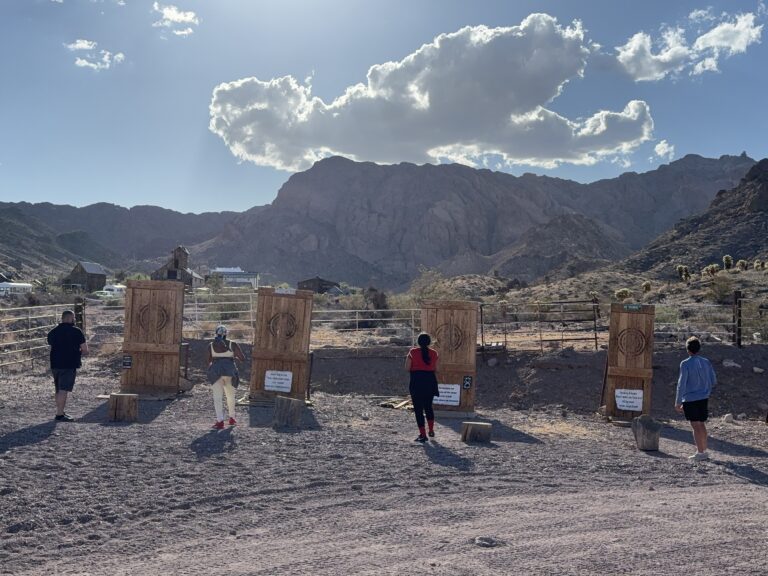 Group axe throwing in Nevada ghost town outdoors, with mountains behind; signs hint at ATV tours and Colorado River adventures.
