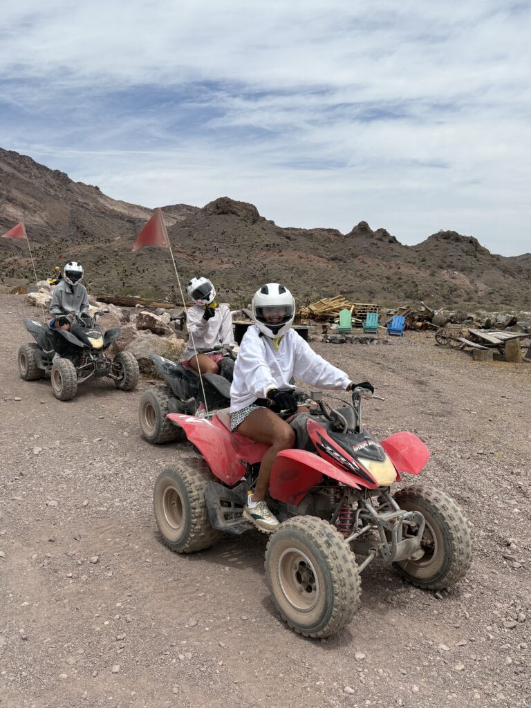 Riders in helmets enjoy an ATV tour on a dusty Nevada trail near Las Vegas, with desert mountains and blue sky in the background.