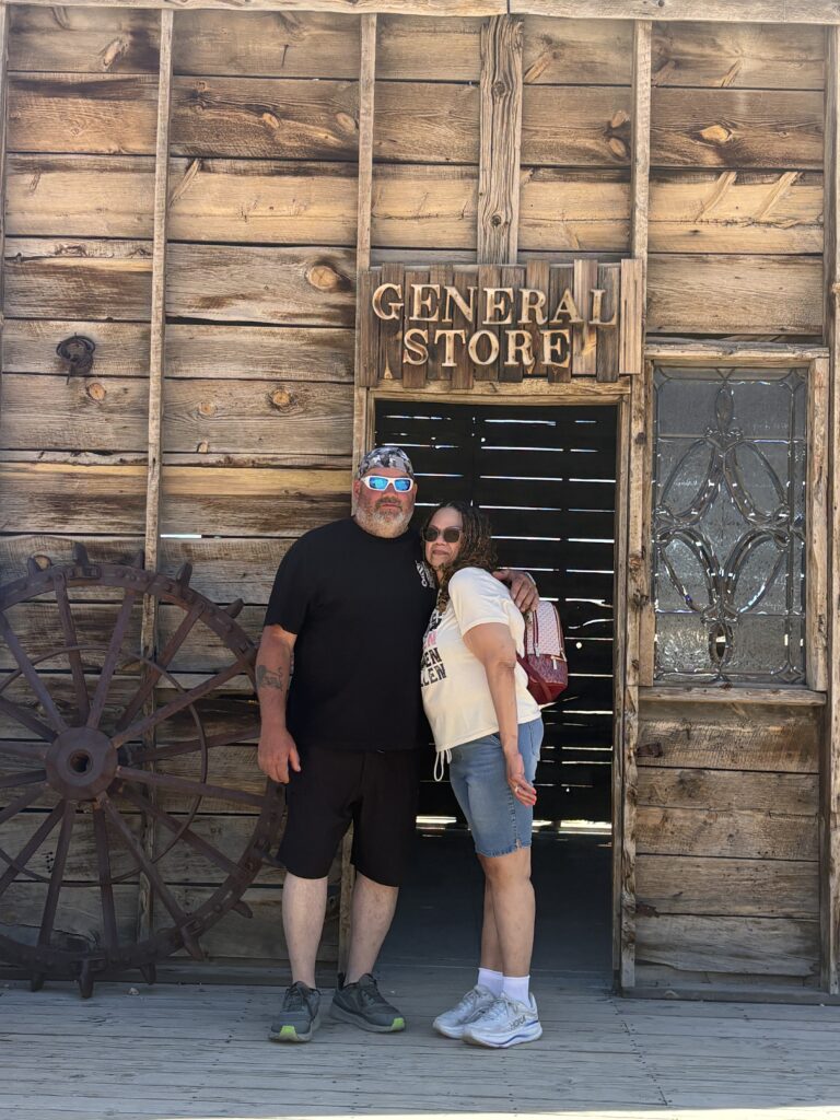 Smiling visitors at a rustic Nevada General Store near Las Vegas, wagon wheel by the wall—perfect Ghost Town sightseeing spot.
