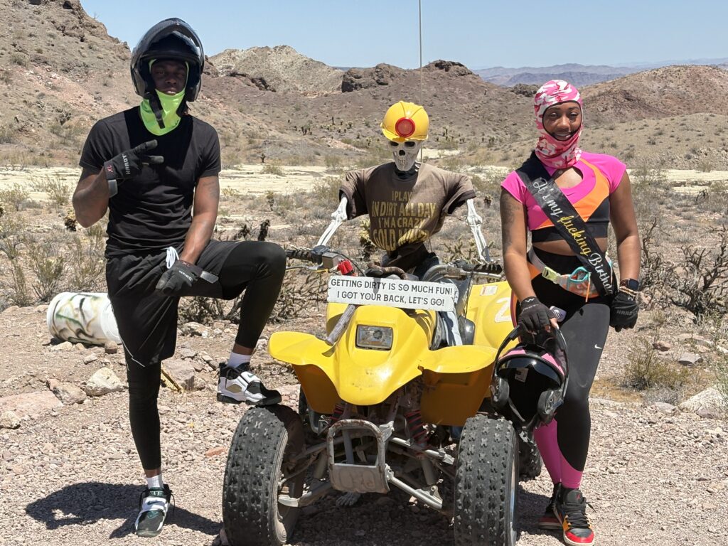Three friends pose near Las Vegas in Nevada desert on a yellow ATV, dressed for an ATV tour with helmets and costumes.