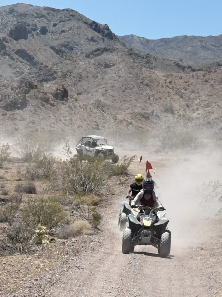 Adventurers on an ATV tour in the Nevada desert near Las Vegas, with a RZR off-road vehicle kicking up dust along rocky desert trails.