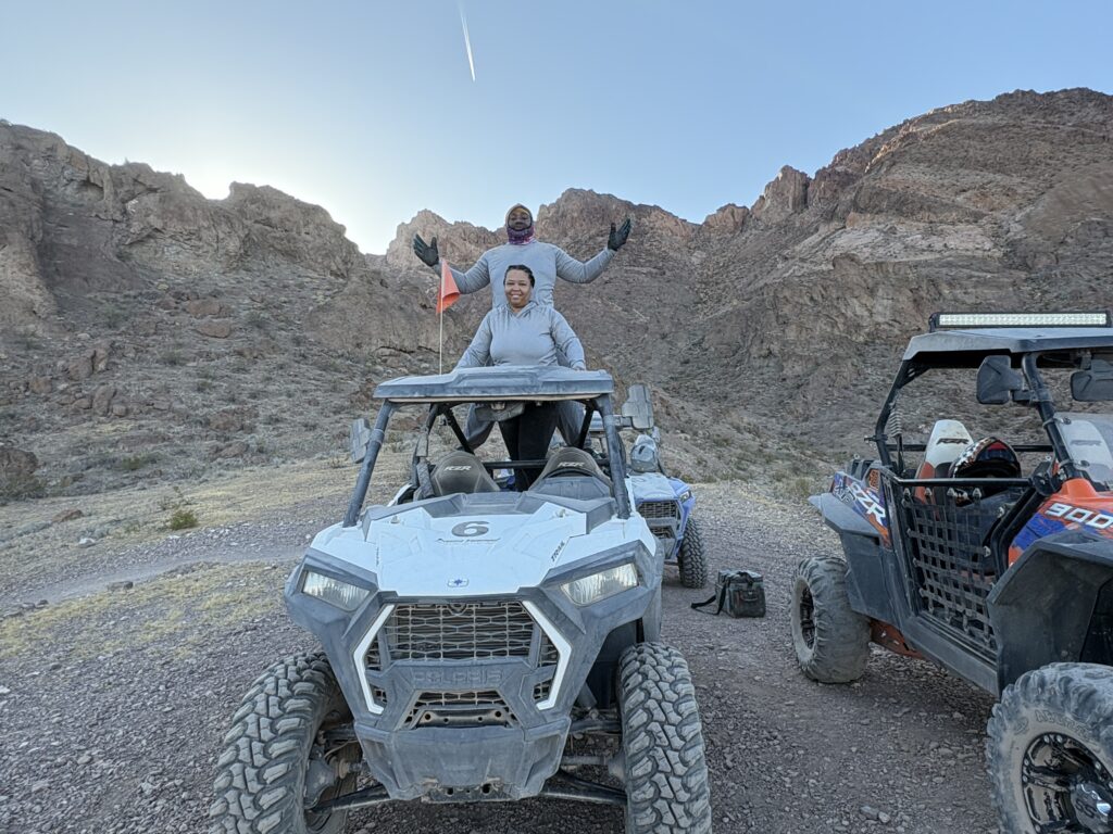 Two smiling people atop a white RZR in Nevada's rocky desert near Las Vegas, enjoying an ATV tour under a clear blue sky.