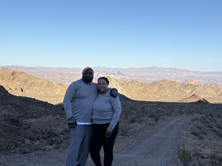 Couple smiling on Nevada desert trail near Las Vegas, mountains behind them, dressed for ATV tours or RZR off-road rides.
