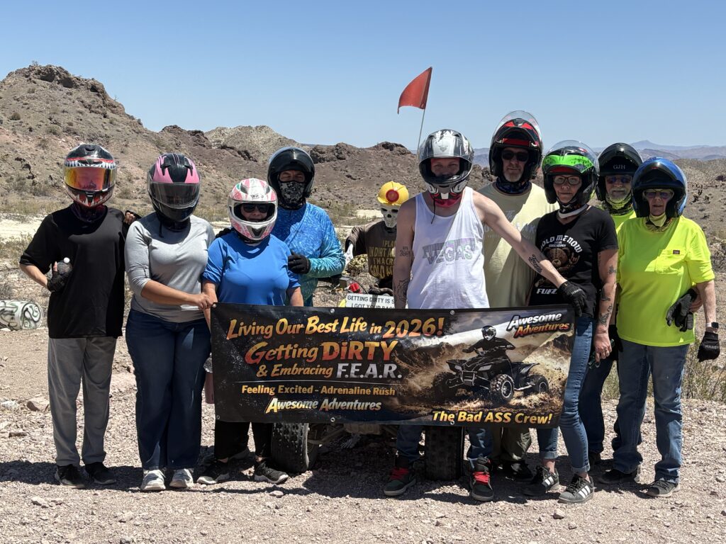 Nine adventurers in helmets on rocky terrain by Las Vegas hold an ATV tour banner, with Nevada mountains and blue sky behind.