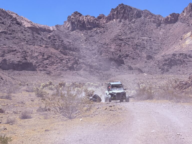 A green RZR ATV speeds down a dusty Nevada desert trail near Las Vegas, with mountains and clear blue sky in the background.