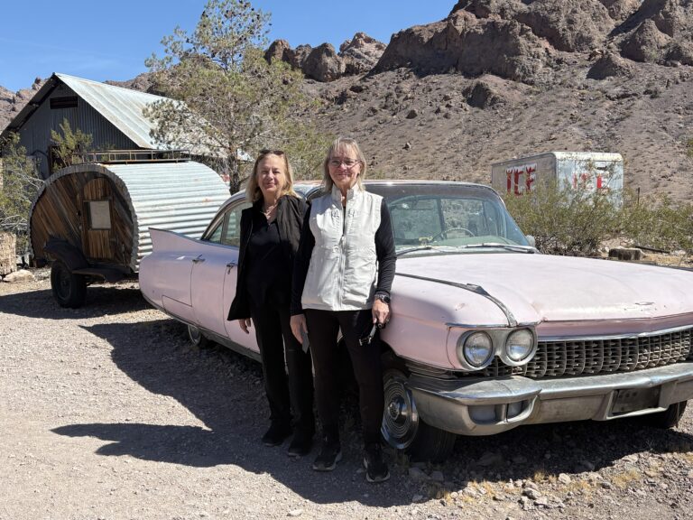 Smiling women by a vintage pink car in a Nevada desert ghost town near Las Vegas, with rocky hills and trailer backdrop.