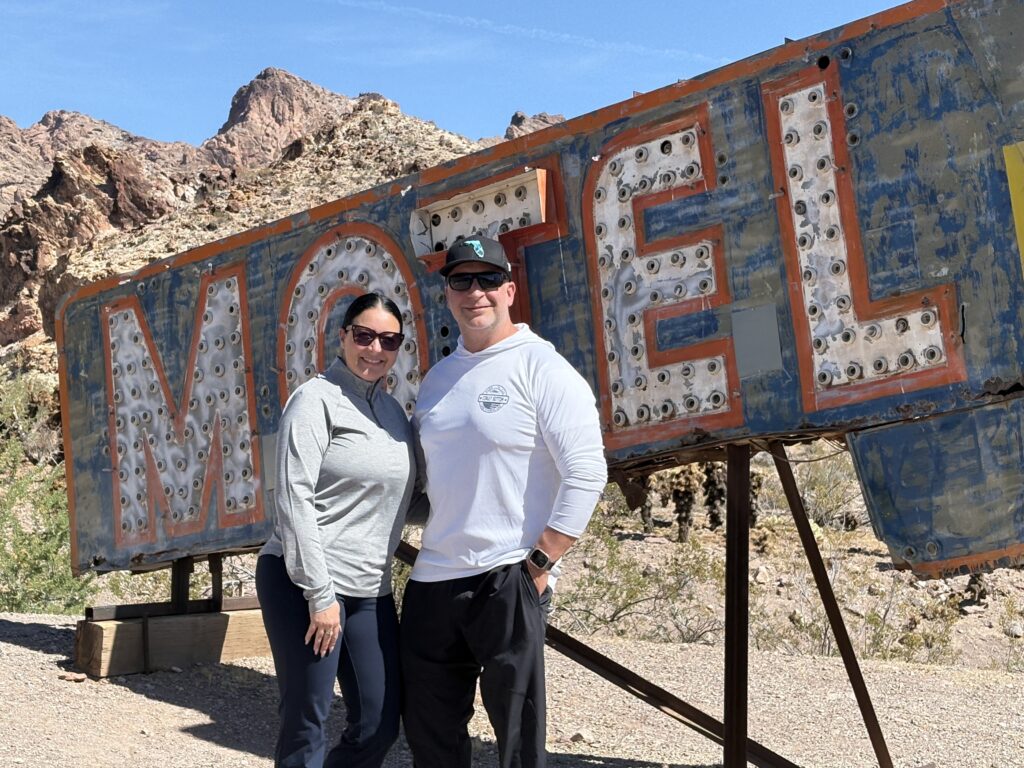 Smiling couple by a vintage MOTEL sign in the Nevada desert near rocky hills—perfect for Vegas trips and Ghost Town sightseeing.