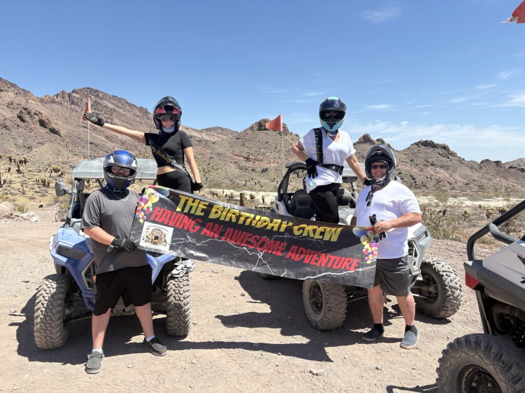 Group with helmets and sunglasses hold a "Birthday Crew" banner by ATVs in Nevada desert near Las Vegas on an ATV tour.
