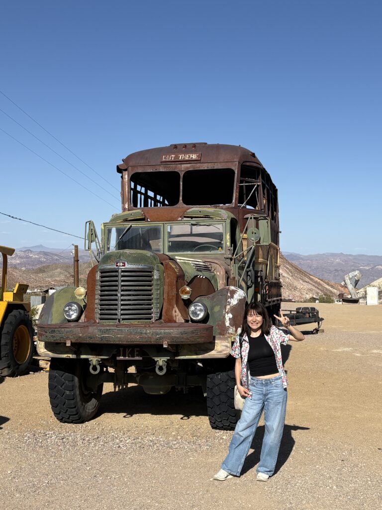 A woman poses in front of a rusty, old military bus in a desert landscape.