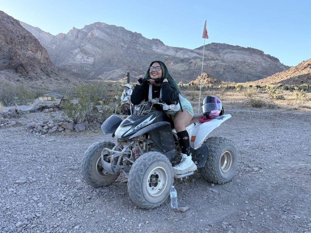 A young woman with green hair and glasses sits on an ATV in a desert landscape, smiling.