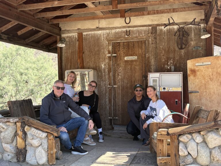 Group smiles on rustic wooden porch in Nevada ghost town near Colorado River, surrounded by vintage decor and stonework.