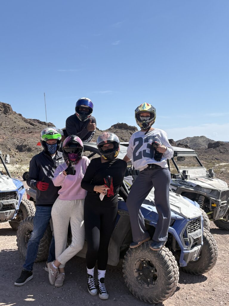Group in helmets and masks by ATVs in Nevada desert, ready for RZR off-road tour near Las Vegas under clear blue sky.