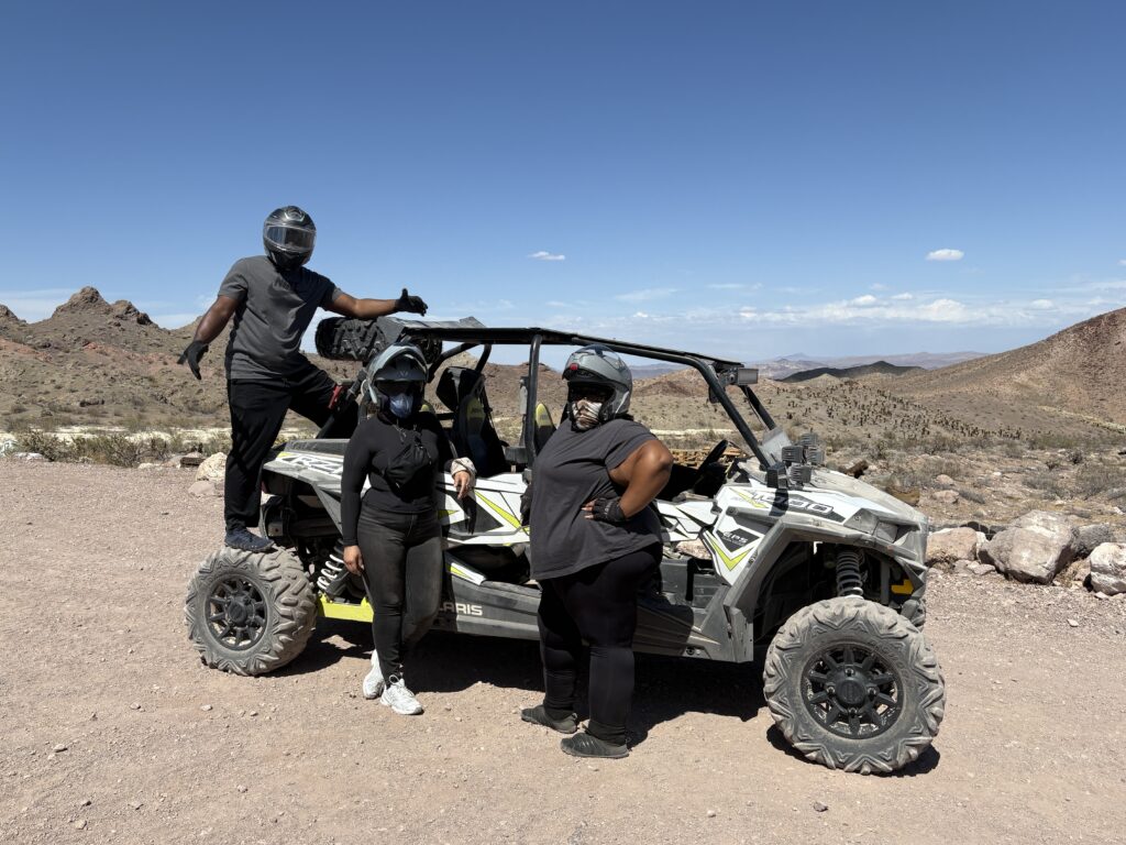 Three riders with helmets on a Polaris RZR during an ATV tour near Las Vegas, Nevada, rocky desert and mountains behind them.