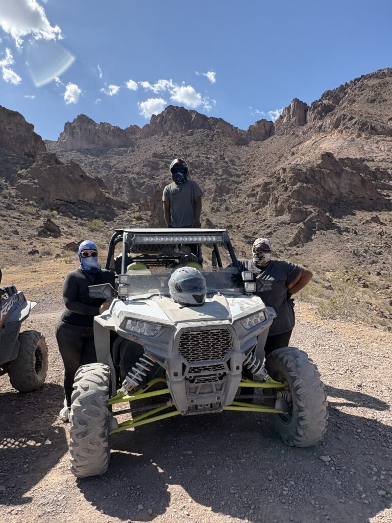 Adventurers in dark clothing with face coverings pose by an ATV in Nevada’s rocky desert near Las Vegas, blue sky and mountains behind.