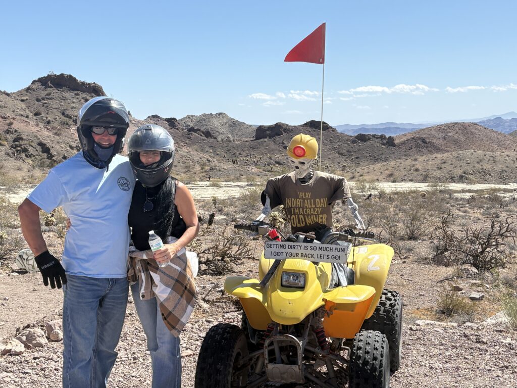 Smiling couple in helmets pose by a yellow ATV and mannequin during an exciting Las Vegas desert ATV tour near the Colorado River.