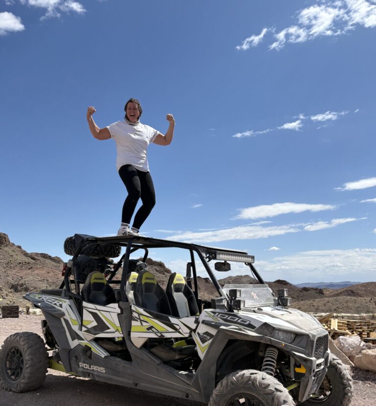 Person in white shirt & black pants celebrates atop a Polaris ATV in Nevada desert near Las Vegas under blue sky—RZR off-road adventure.
