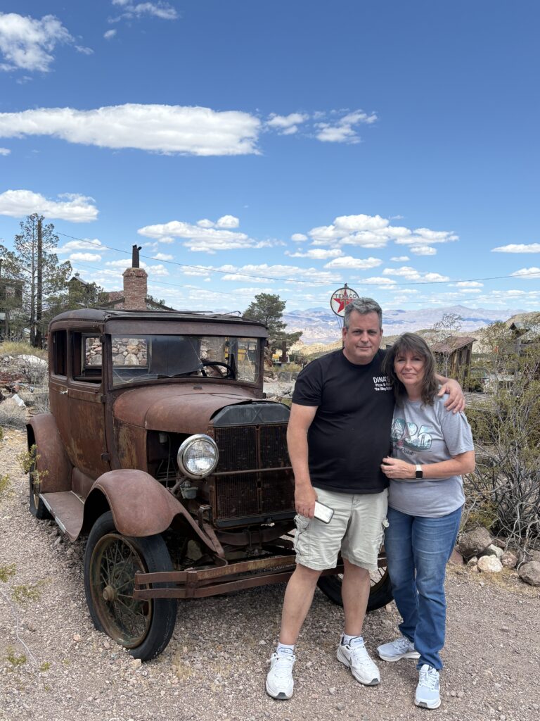 A smiling couple poses by a rusty vintage car in a Nevada ghost town near Las Vegas, perfect for sightseeing and ATV tours.