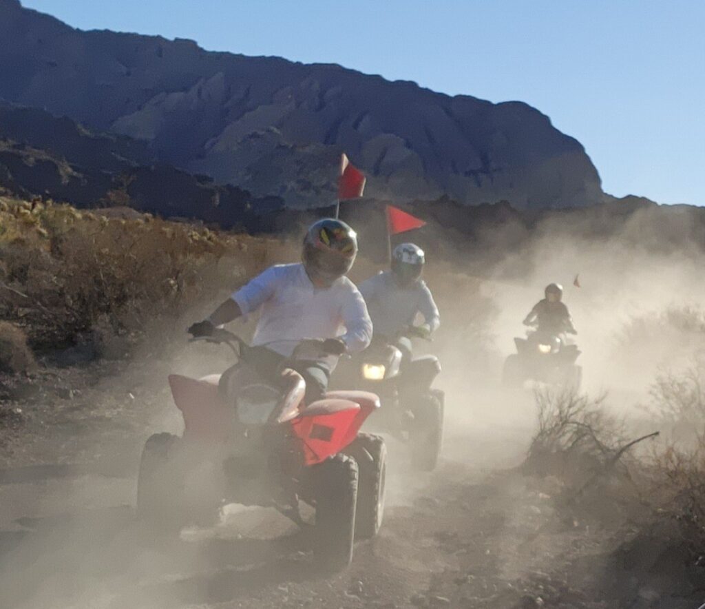Riders on red ATVs speed along a dusty Nevada trail near Las Vegas, with mountains and red flags, under a clear blue sky.