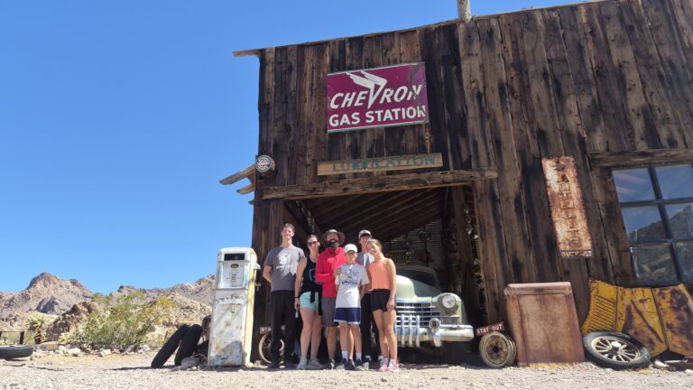 Group smiles at rustic wooden building with vintage Chevron sign in Nevada’s desert near Las Vegas, perfect for Ghost Town sightseeing.