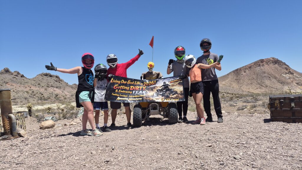 Group in helmets pose with a quad bike and banner on a Nevada ATV tour near Las Vegas, rocky hills and blue sky behind them.