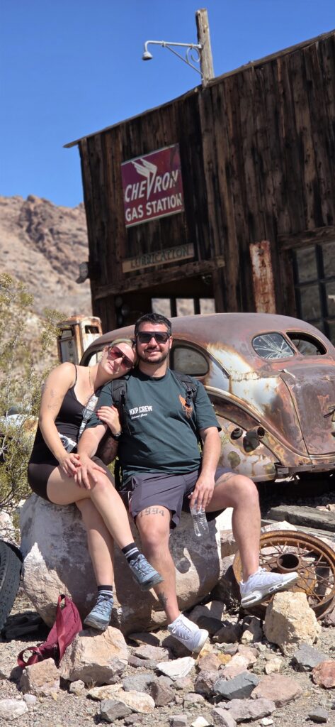 A smiling couple sits on a rock by a rusty car at a vintage Chevron gas station near Las Vegas on a Nevada ghost town tour.