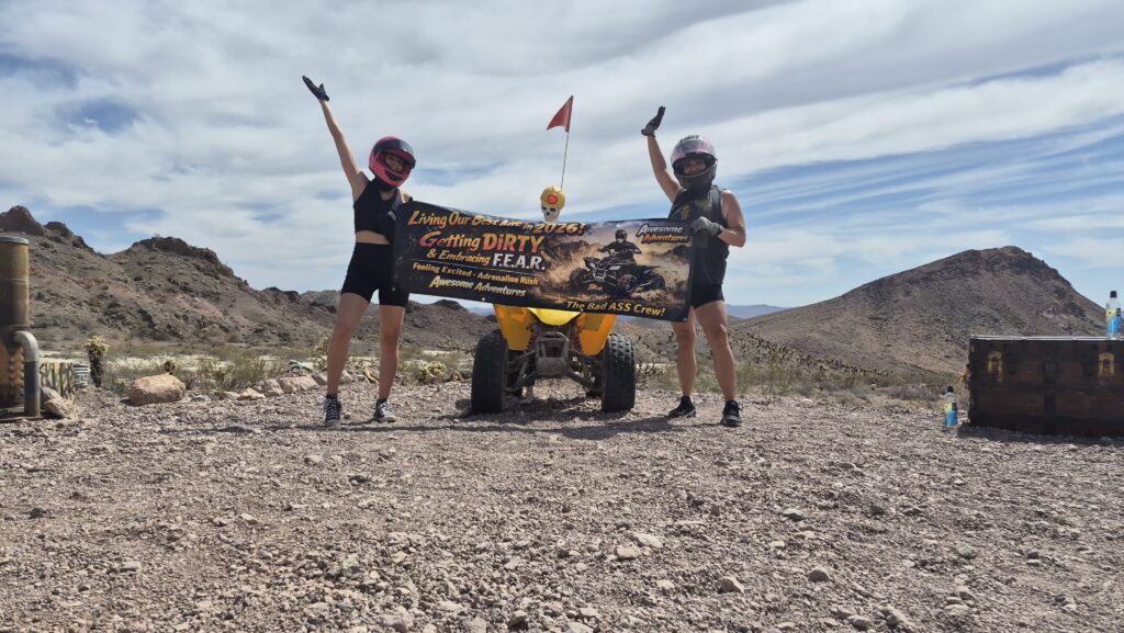 Two people in helmets pose by a yellow ATV in the Nevada desert, holding a “Getting Dirty” banner on an ATV tour near Las Vegas.