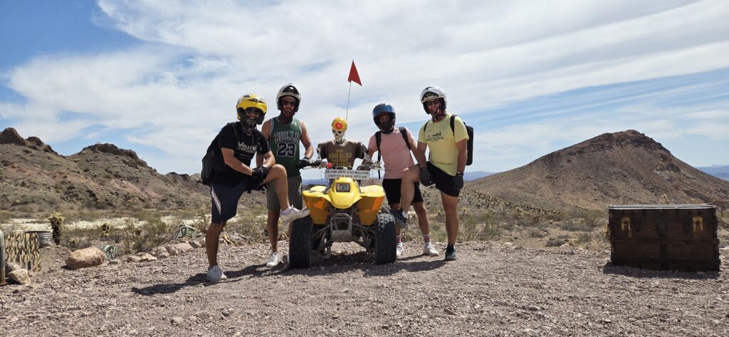 Group in helmets poses with yellow ATV on Nevada desert tour near Las Vegas; mountains and wooden chest complete the scene.