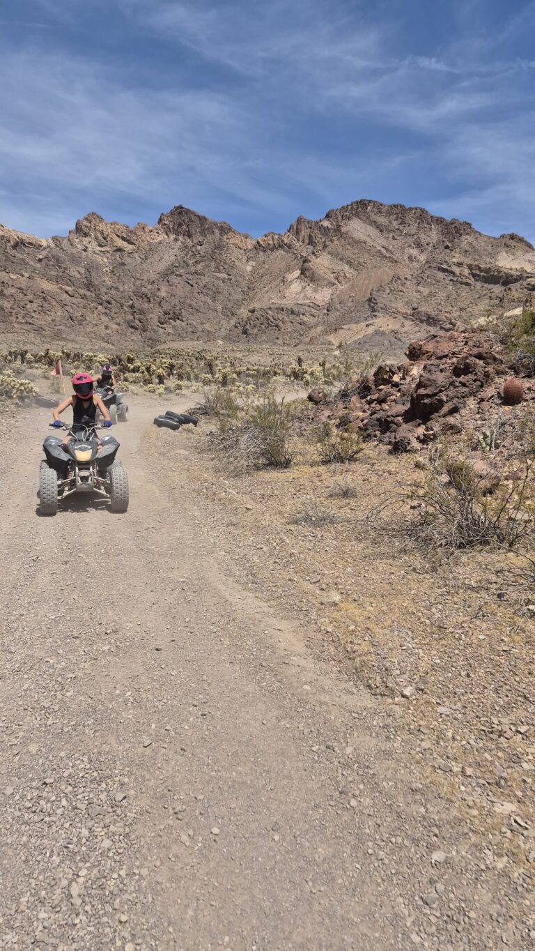 Adventurer in red helmet on ATV rides a dusty Nevada desert trail near Las Vegas, with mountains and blue sky—perfect for ATV tours.