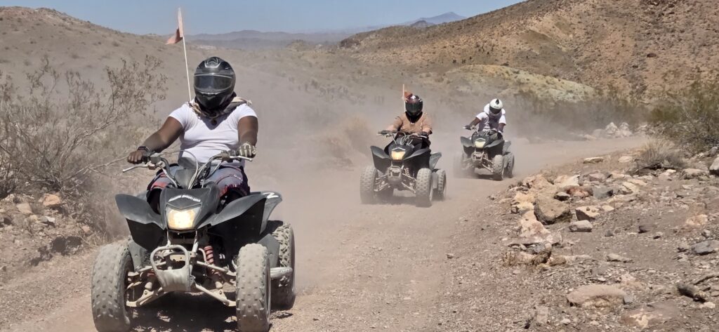 Adventurers on an ATV tour ride dusty trails near Las Vegas, Nevada, surrounded by rocky desert hills under a clear sky.