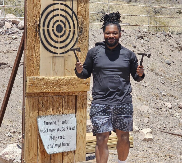Smiling man at a Las Vegas axe throwing site, holding two axes by a target with funny instructions, ready for adventure fun.