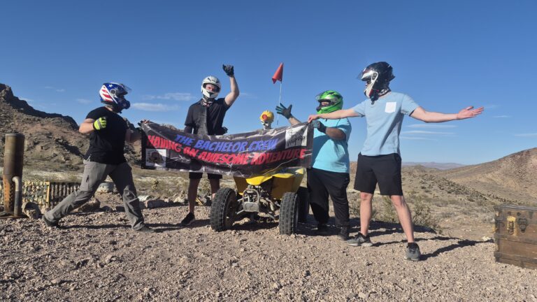 Four helmeted friends with ATV banner in Nevada desert near Las Vegas, perfect for RZR off-road tours & Colorado River adventure.