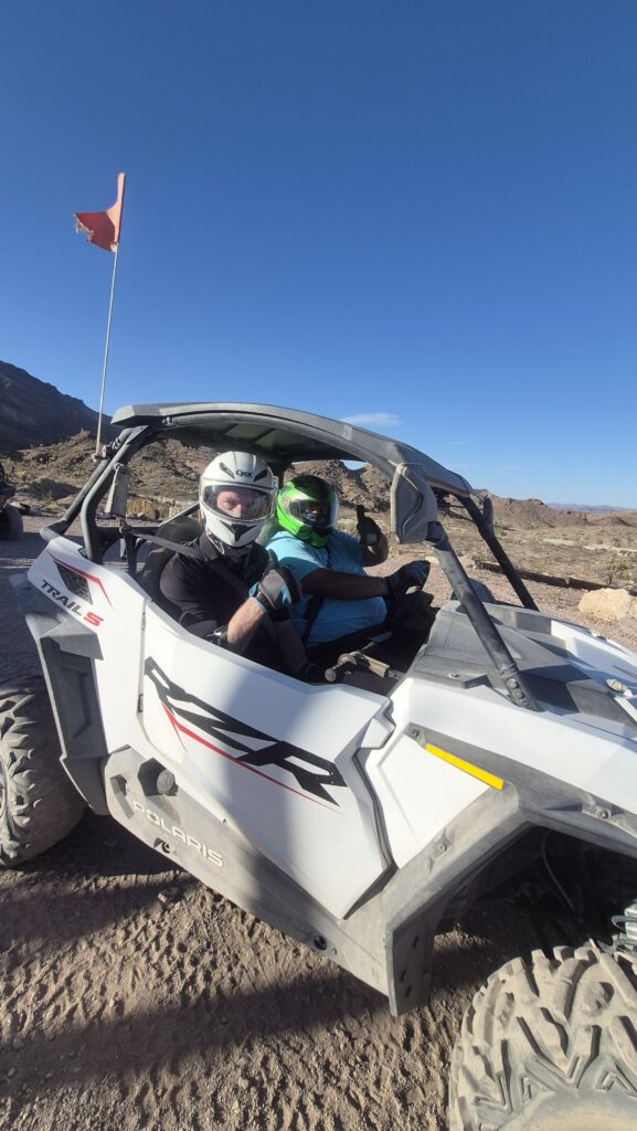 Two riders in helmets sit in a white RZR with an orange flag on a rocky Nevada desert trail near Las Vegas, perfect for ATV tours.