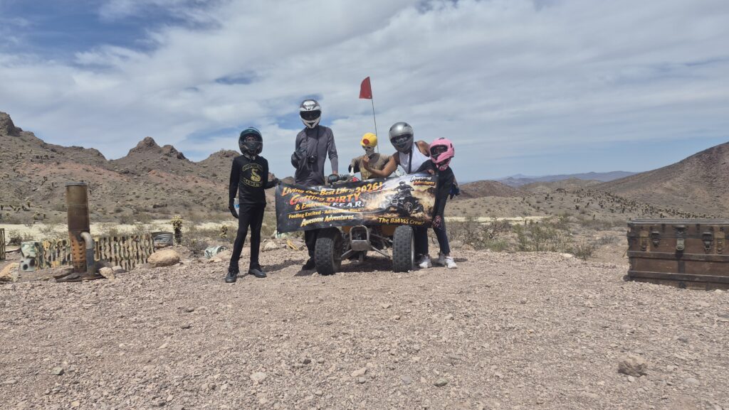 Group in helmets holds dirt road adventure banner by ATV, Nevada desert. Mountains, cloudy sky, and treasure chest near Las Vegas.