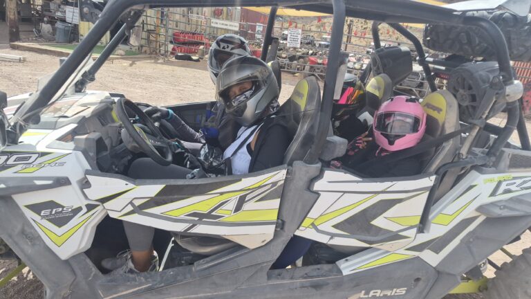 Four people in helmets sit in a RZR for a Nevada ATV tour near Las Vegas, with ATVs and desert gear in the background.