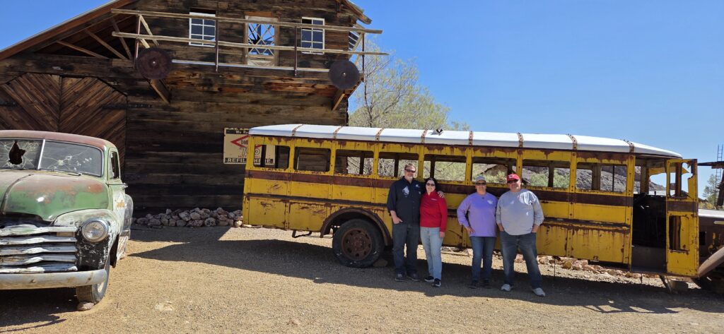 Four people smile by a rusty yellow bus in a Nevada ghost town, with an old green truck—perfect for Ghost Town sightseeing tours.