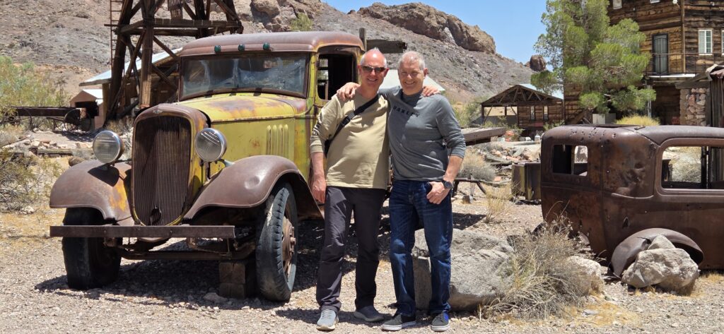 Two men smile arm-in-arm by a rusty yellow truck in a Nevada ghost town near Las Vegas—perfect for ATV and RZR sightseeing tours.
